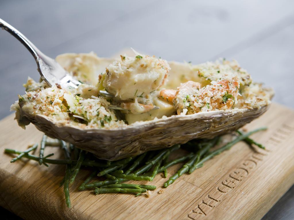 Montcalm Brewery Townhouse Hotel Лондон Екстериор снимка The photo shows a large oyster shell filled with a mixture that appears to include seafood, herbs, and breadcrumbs. The dish is presented on a wooden board with some greenery, possibly a type of seaweed or garnishing. A fork is placed beside the oyst