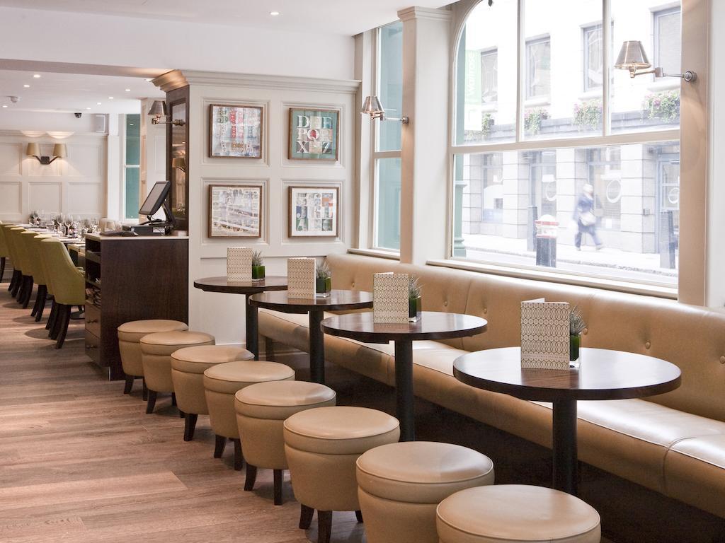 Montcalm Brewery Townhouse Hotel Лондон Екстериор снимка The photo displays a modern café or restaurant interior. There is a cozy seating area featuring light-colored, upholstered benches along the wall, accompanied by small round tables. The tables are paired with circular stools. The decor includes frame
