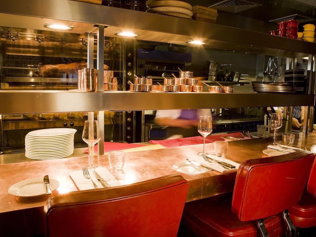 Montcalm Brewery Townhouse Hotel Лондон Екстериор снимка The photo shows a restaurant interior featuring a bar or counter area. There are red chairs arranged around a copper-topped table where dining utensils and plates are set. Above the counter, there are shelves displaying various cooking pots and kitch