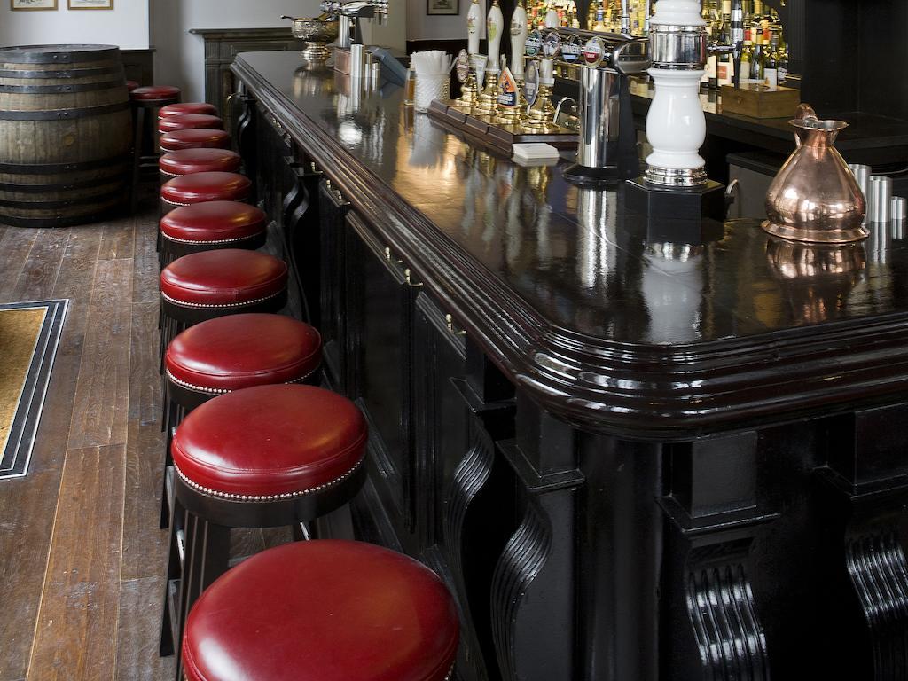 Montcalm Brewery Townhouse Hotel Лондон Екстериор снимка The photo shows a bar setting with a long, polished dark wooden bar counter. Attached to the counter are several bar stools with red leather seats. Behind the bar, there are various bottles, suggesting a well-stocked bar. The flooring appears to be w